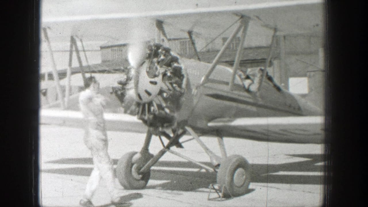 A vintage open-cockpit biplane rests on a paved airfield in the supplied Wyoming 1939 clip, with a man standing near the engine and a person visible in the cockpit.