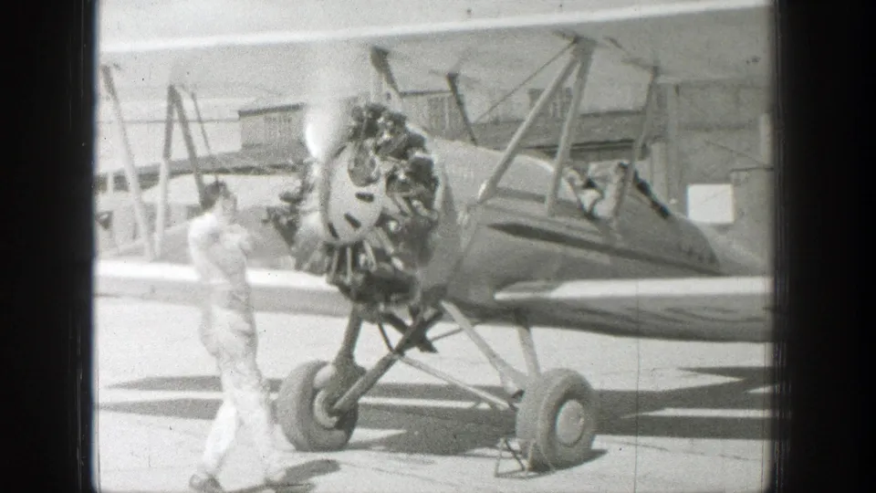 A vintage open-cockpit biplane rests on a paved airfield in the supplied Wyoming 1939 clip, with a man standing near the engine and a person visible in the cockpit.