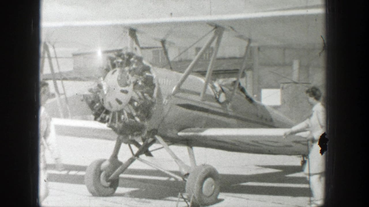 Biplane fills the frame as a person sits in the cockpit and another stands near the wing.