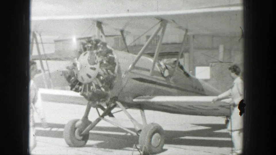 The biplane is seen closer on the tarmac, with its radial engine and struts prominent, a cockpit occupant visible, and another person standing near the right side of the aircraft.