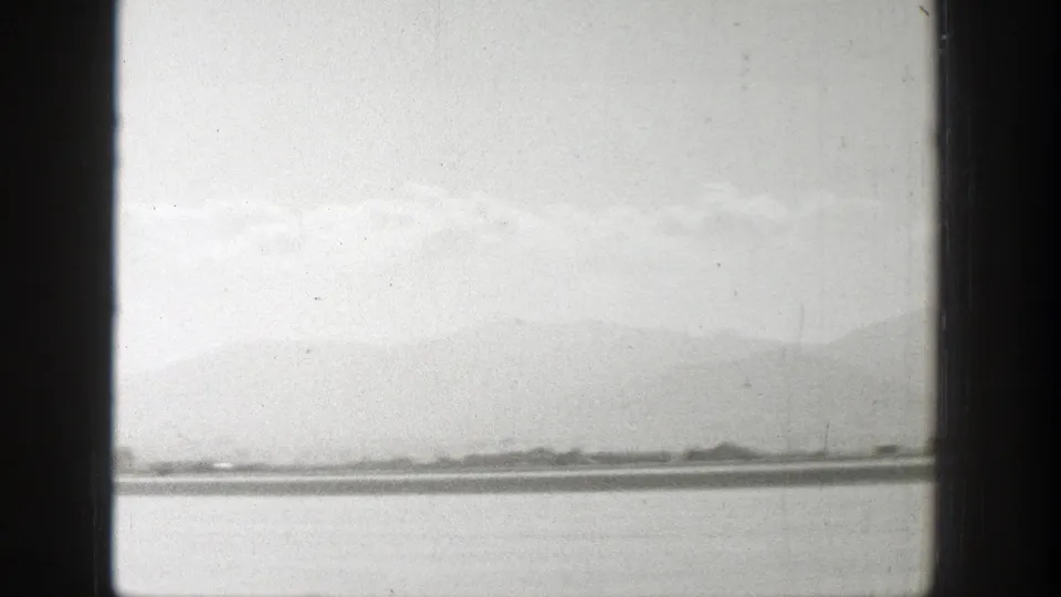 1939 Wyoming home-movie view of a distant silver plane on a runway, framed by dark vertical window bars and mountains beyond.
