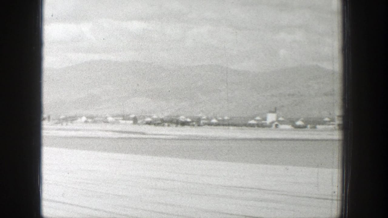 Aircraft-window view over a pale valley with a distant town and mountains beyond.