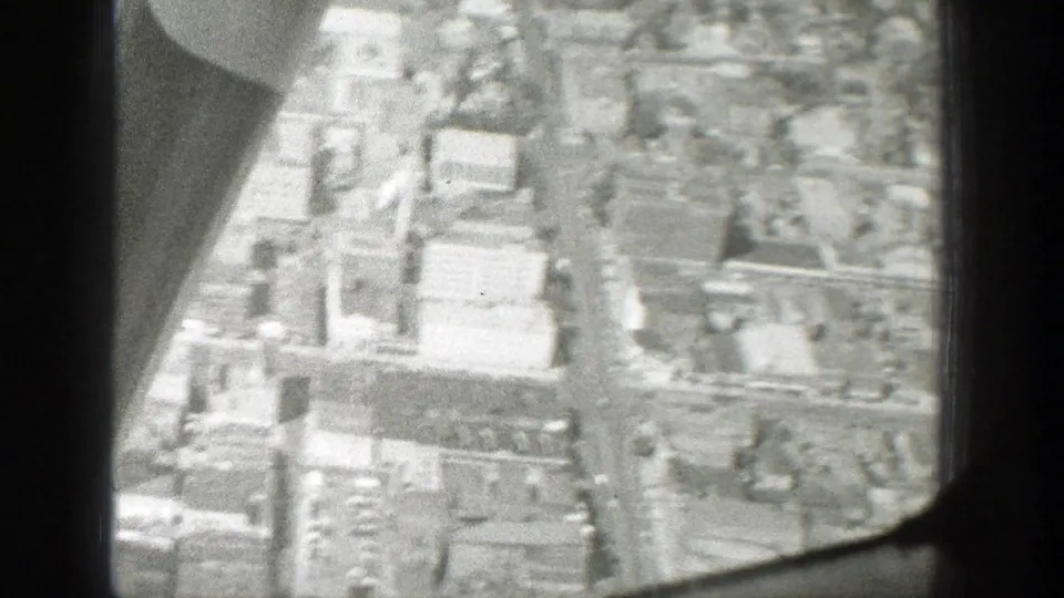 Closer 1939 aerial home-movie view of urban blocks and large pale buildings, with part of the aircraft visible at left.