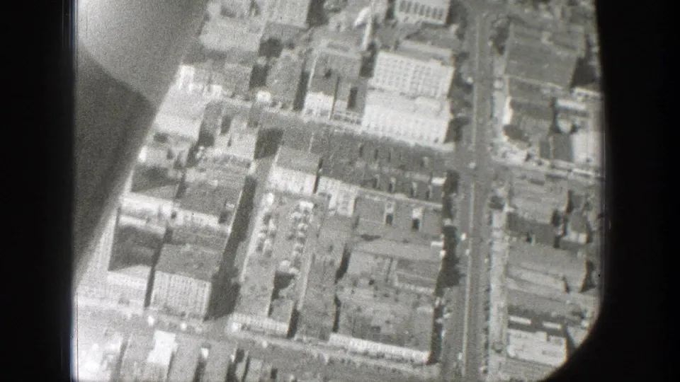 Black-and-white 8mm frame showing dense city blocks and streets from above through the side of an airplane window.