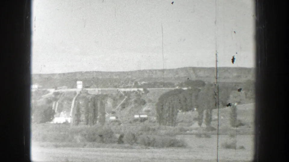 Distant view of a dam and hillside landscape, with small-gauge film marks visible in the frame.