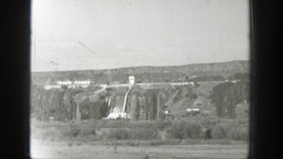 Distant dam view with Idaho Power signage visible on the hillside, framed by the aircraft window edge.