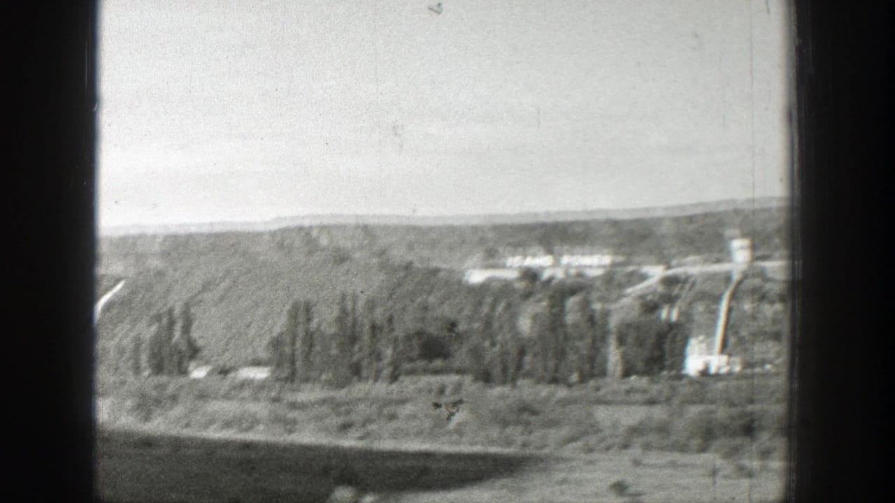 Distant dam or hydroelectric works in a wooded Western valley, with the view partially blocked by a dark vertical window frame.