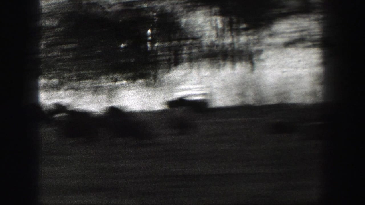 High-contrast monochrome frame from a moving vehicle, with roadside trees and terrain blurred into dark streaks under a bright sky band.