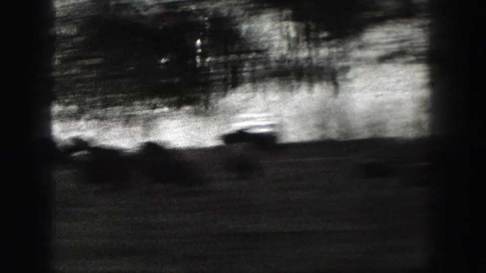 High-contrast monochrome frame from a moving vehicle, with roadside trees and terrain blurred into dark streaks under a bright sky band.