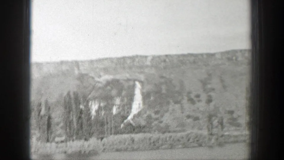 1939 Wyoming landscape frame showing a pale hillside, a bright winding road or snow-like streak, and sparse trees seen from a moving vehicle.