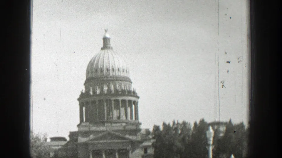 Black-and-white Wyoming home-movie frame of a large domed civic building, likely the Wyoming State Capitol, partially framed by trees.
