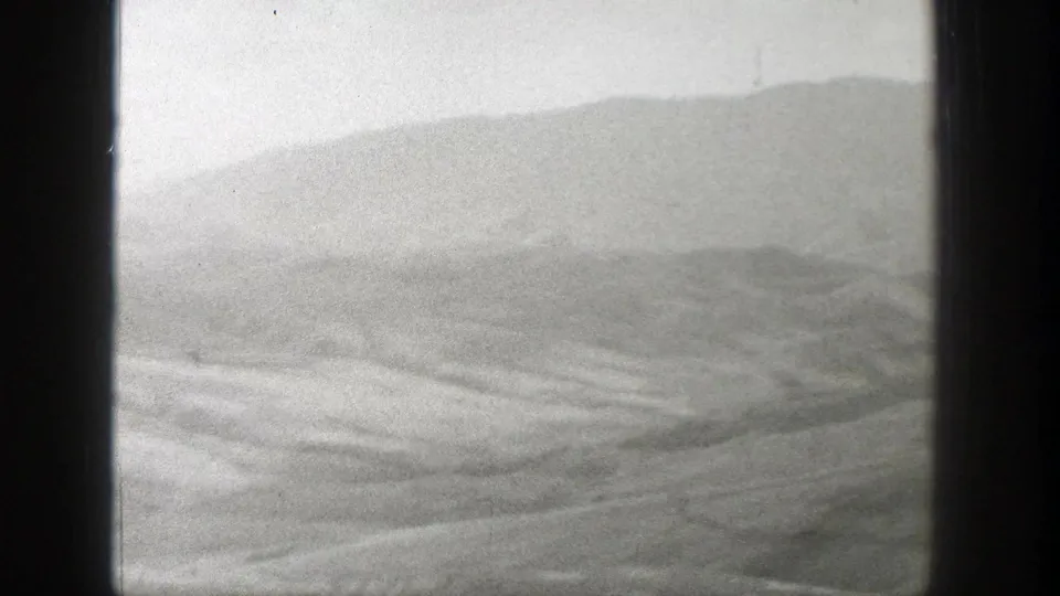 1939 Wyoming home-movie frame with a barren foreground, layered low ridges, and a muted mountain slope in the distance.