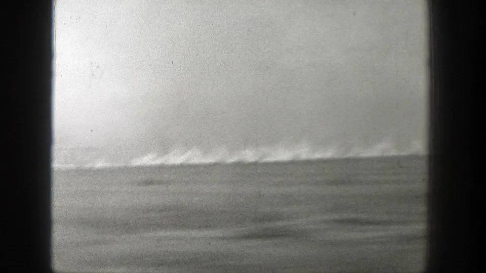 1939 Wyoming home-movie frame with a low line of flame or smoke crossing a flat grass field beneath a washed-out sky.