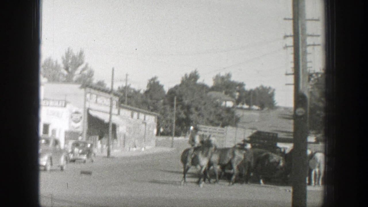 Mounted rider leading horses past a small-town street corner with storefronts and cars.