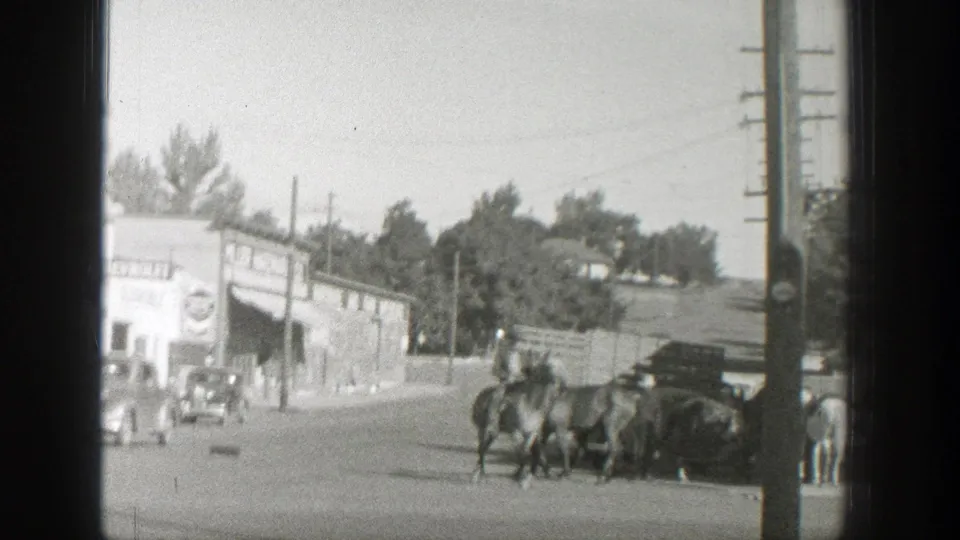 In 1939 Wyoming home-movie footage, a rider leads horses past storefronts, utility poles, and parked vintage automobiles on a bright street.