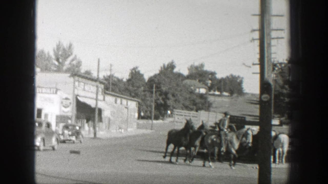 The rider leads horses across a Wyoming town street with a Chevrolet-marked storefront, vintage automobiles, and utility poles in view.