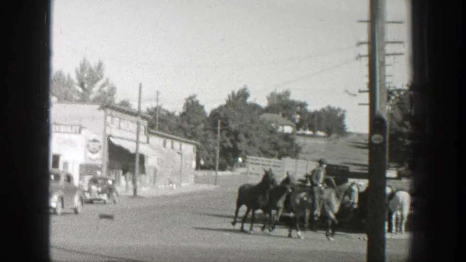 The rider leads horses across a Wyoming town street with a Chevrolet-marked storefront, vintage automobiles, and utility poles in view.