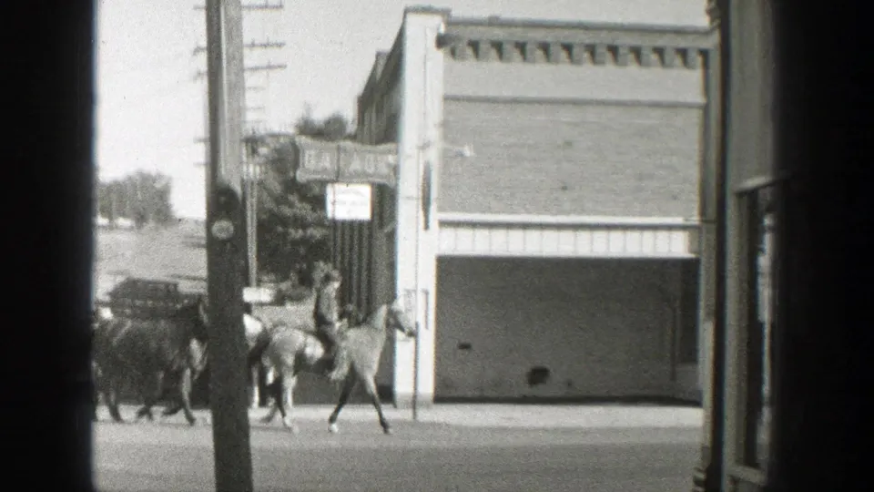 A rider and led horses pass a prominent storefront facade in 1939 Wyoming, with a parked car, sidewalk, and utility pole visible.