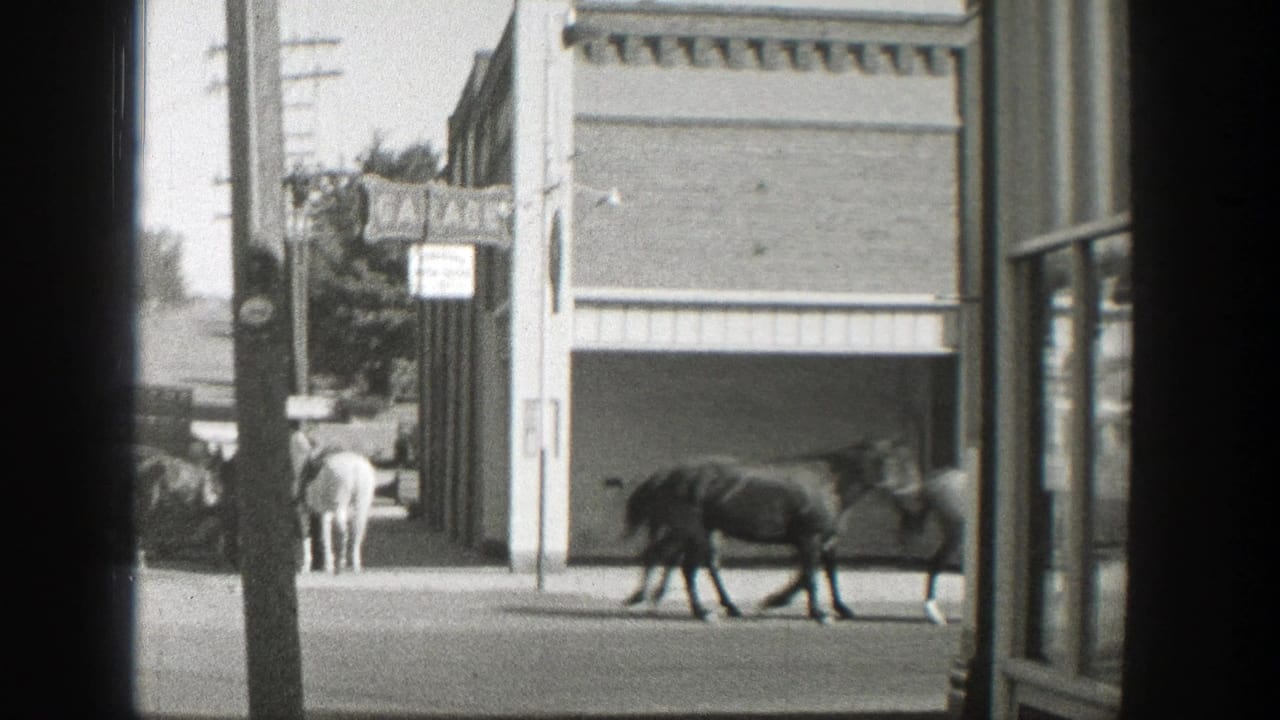 1939 Wyoming home-movie frame showing mounted riders and horses passing storefronts, automobiles, utility poles, and a Coca-Cola sign.