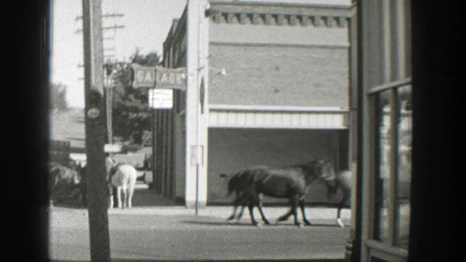 1939 Wyoming home-movie frame showing mounted riders and horses passing storefronts, automobiles, utility poles, and a Coca-Cola sign.