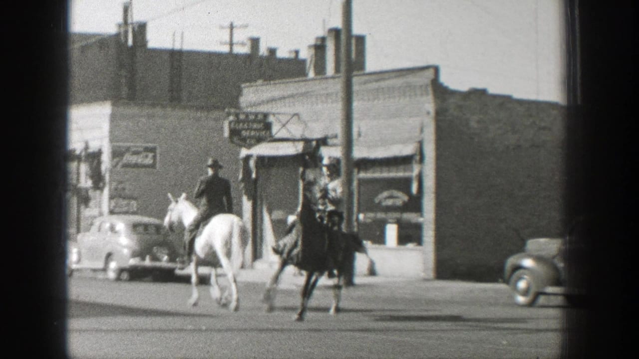 Two cowboy-hatted riders on light and dark horses cross a Wyoming town street in 1939, with storefront signs and cars behind them.