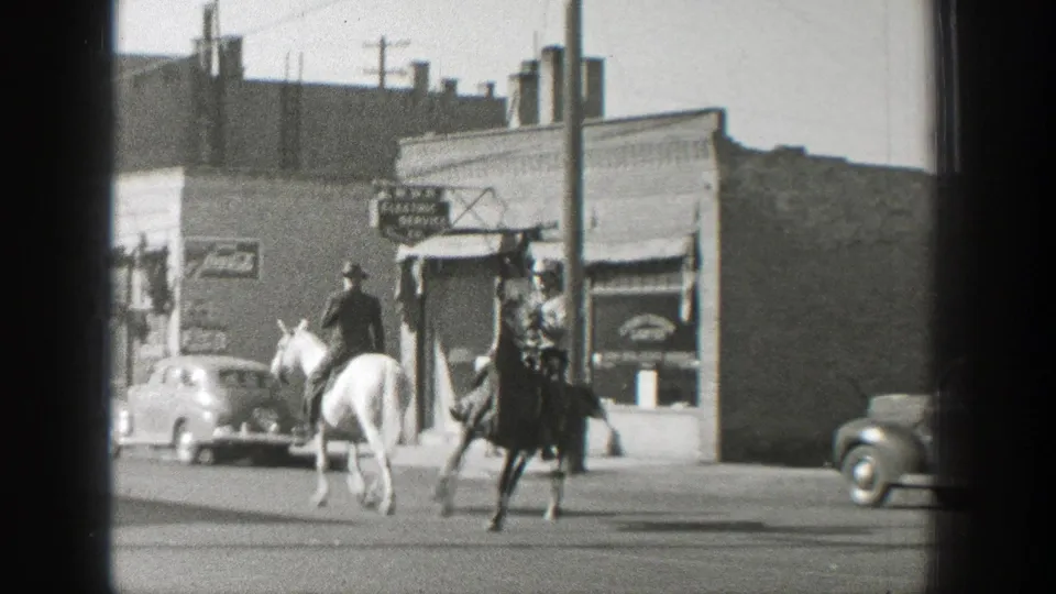 Two cowboy-hatted riders on light and dark horses cross a Wyoming town street in 1939, with storefront signs and cars behind them.