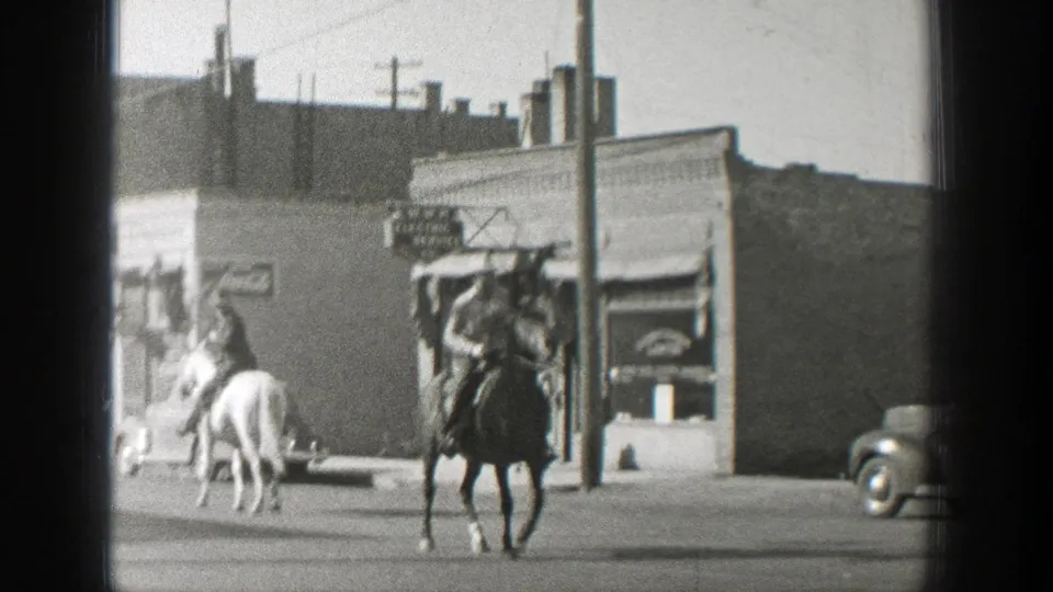 A rider on a dark horse approaches the camera on a 1939 Wyoming street as another horse turns near storefronts and a parked car.