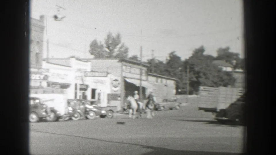 A Wyoming small town street scene shows horseback riders near vintage cars and a Chevrolet storefront in black-and-white 8mm footage.