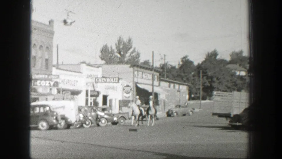 The frame shows riders on horseback near 1930s automobiles, storefront signs, and low buildings on a Wyoming town street.