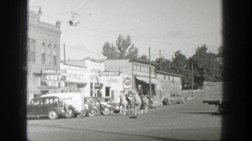 Horseback riders appear near vintage cars and storefronts in a 1939 Wyoming small town street scene.