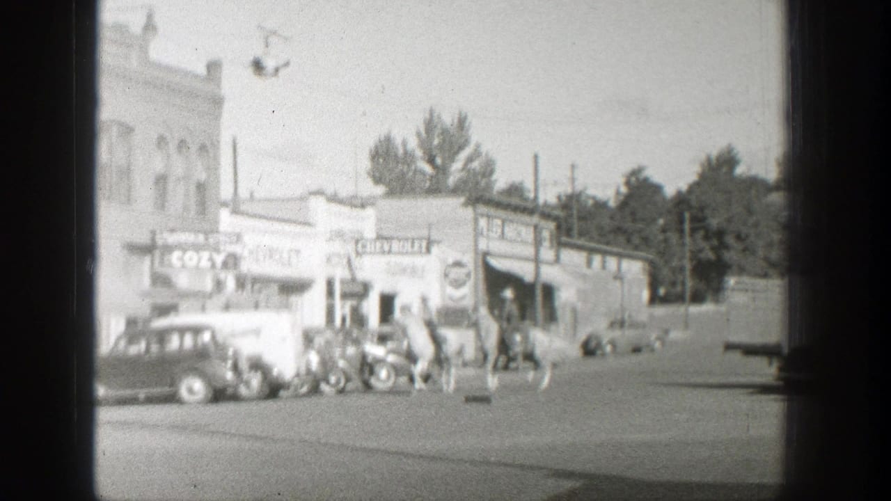 Horseback riders and parked vintage cars on a small-town street.