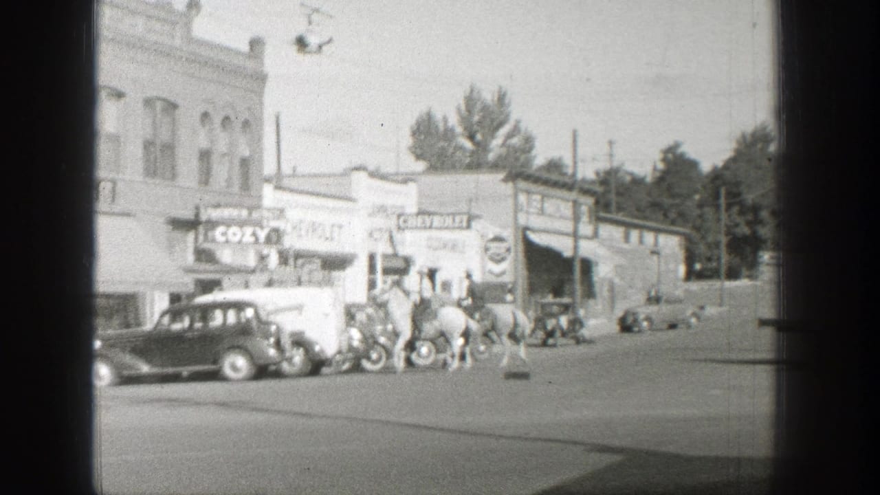 Horseback riders travel along a Wyoming town street in 1939 beside parked 1930s cars and commercial signs.