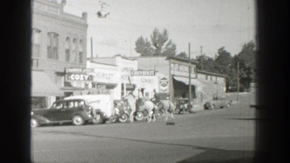 Horseback riders travel along a Wyoming town street in 1939 beside parked 1930s cars and commercial signs.