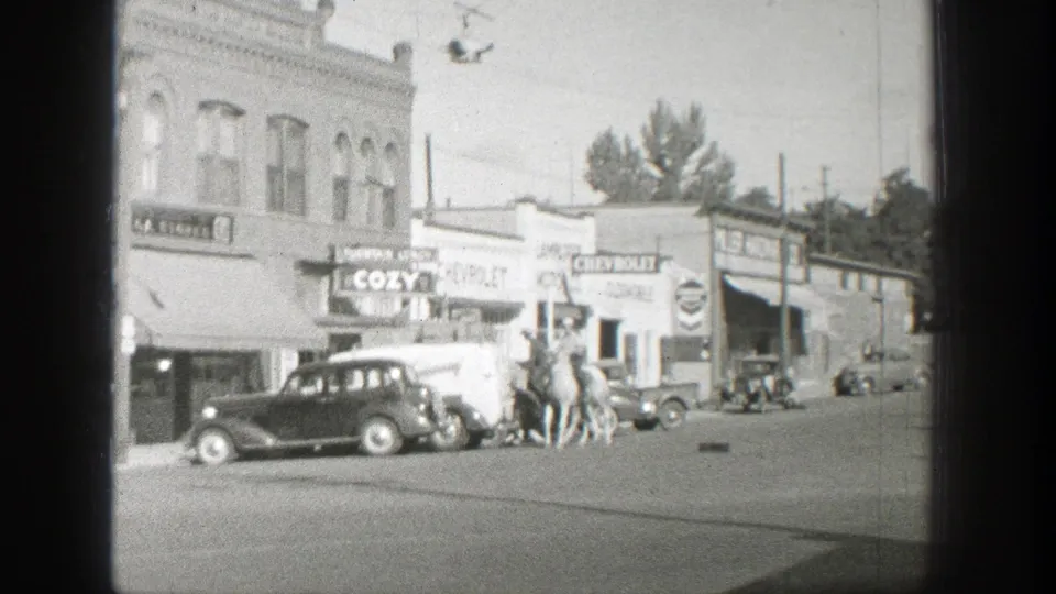 Black-and-white 8mm frame of two riders near vintage automobiles on a Wyoming commercial street, 1939.
