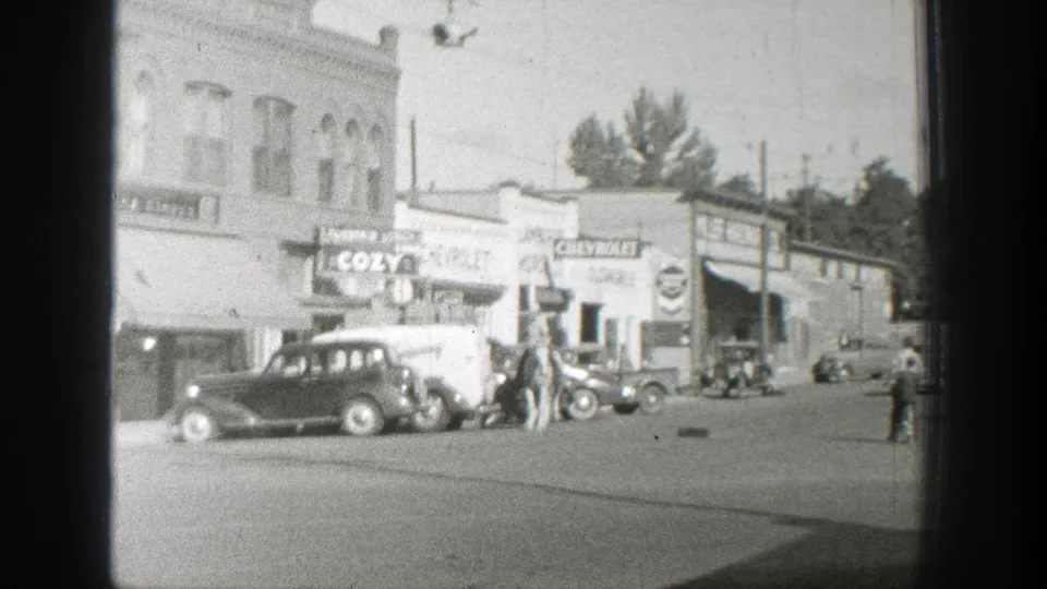 1939 Wyoming home-movie frame showing horseback riders among parked cars, storefront signs, and utility poles.