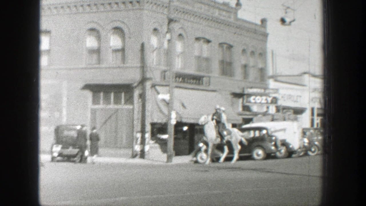Two men ride horses past parked cars and brick storefronts on a town street.