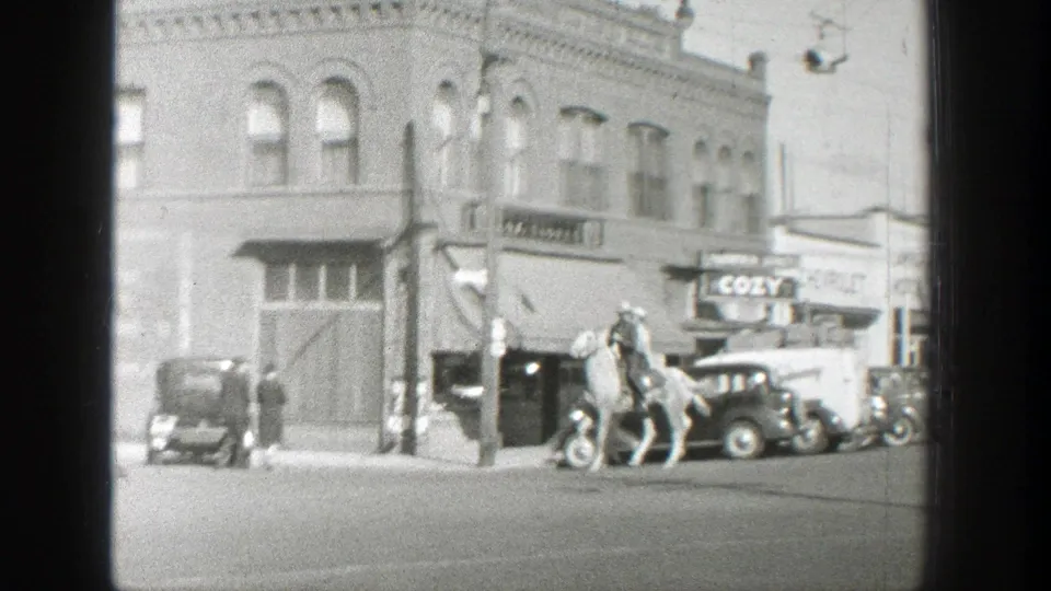 In 1939 Wyoming home-movie footage, two mounted men pass parked automobiles near brick storefronts and a COZY sign.