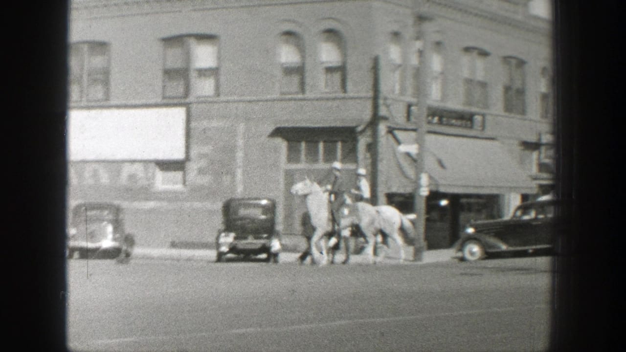 Two riders on horses cross a Wyoming town street in 1939, passing a row of parked cars and commercial building fronts.