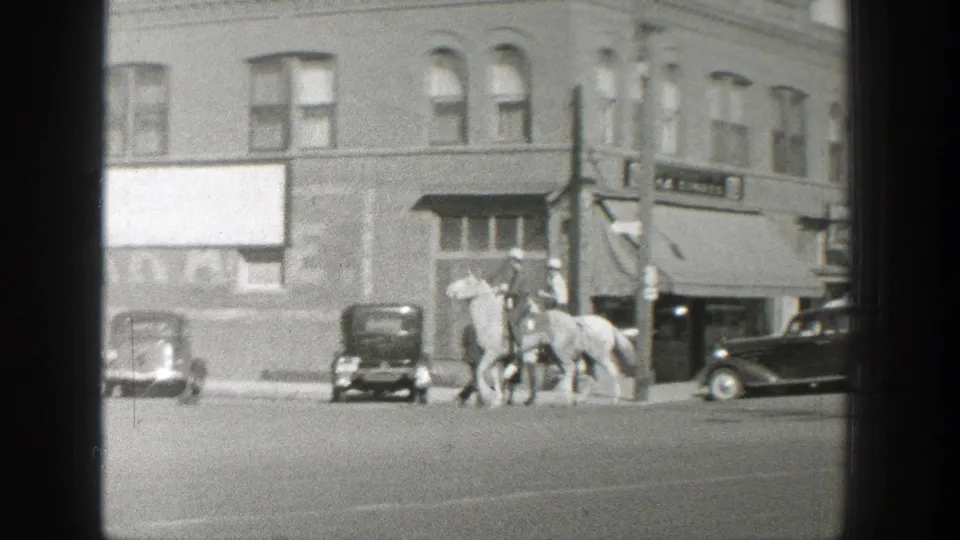 Two riders on horses cross a Wyoming town street in 1939, passing a row of parked cars and commercial building fronts.