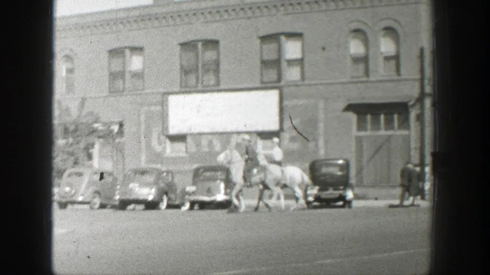 Mounted men move past a row of vintage cars beneath a large signboard on a 1939 Wyoming commercial street.