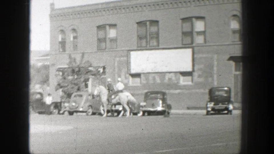 Two horse riders continue along the town street, with parked cars, storefronts, a signboard, and a curbside figure visible.