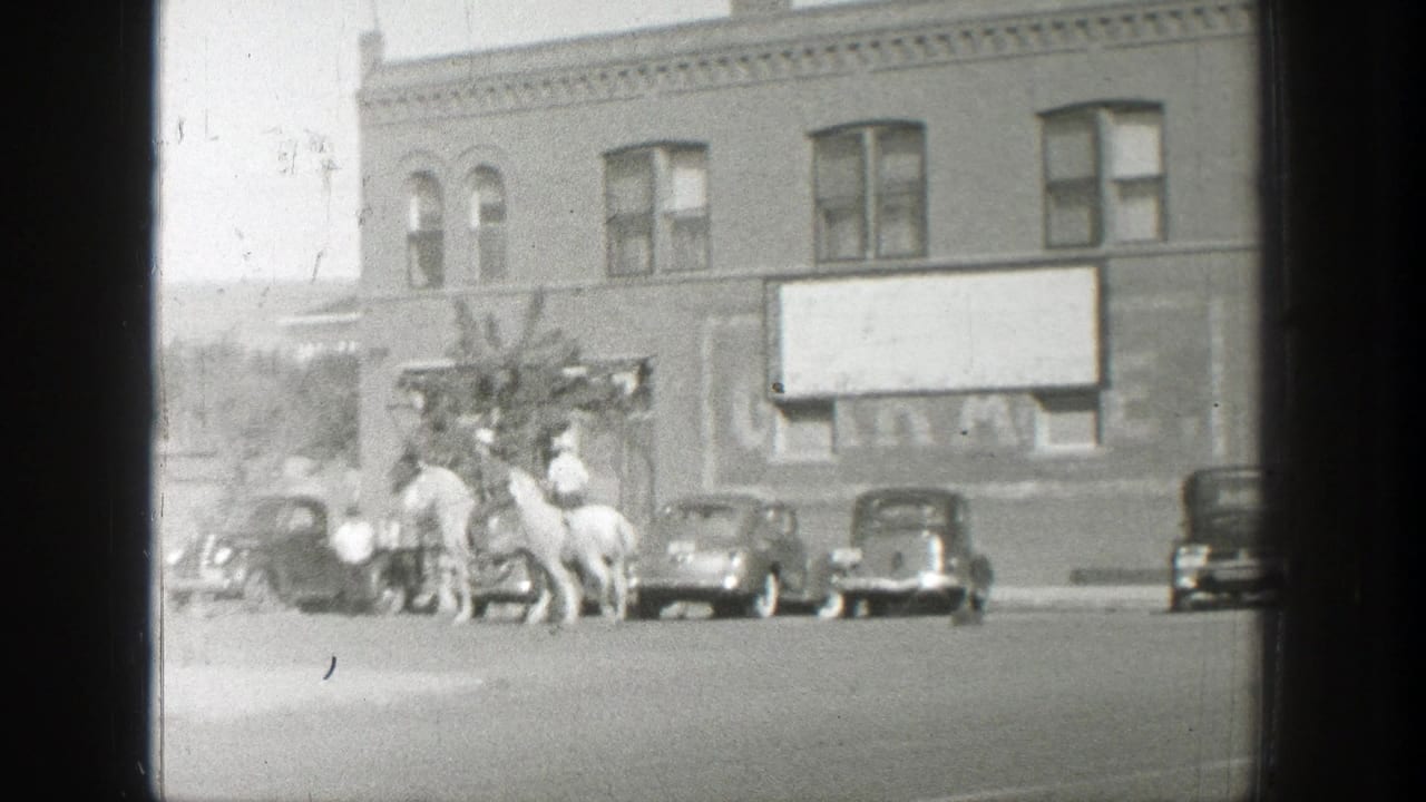 Horse riders and parked cars outside a two-story brick storefront.