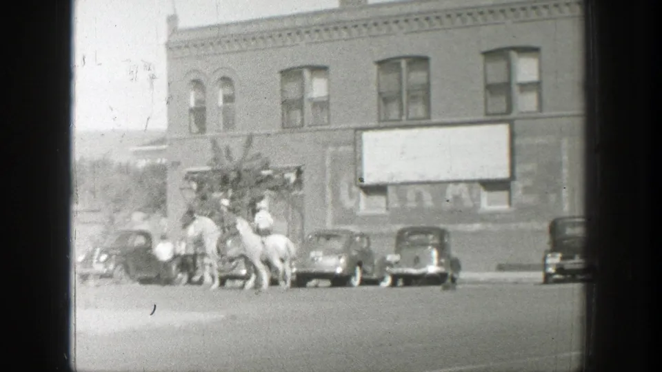 1939 Wyoming home-movie frame with men on horseback near parked automobiles and a brick commercial building.
