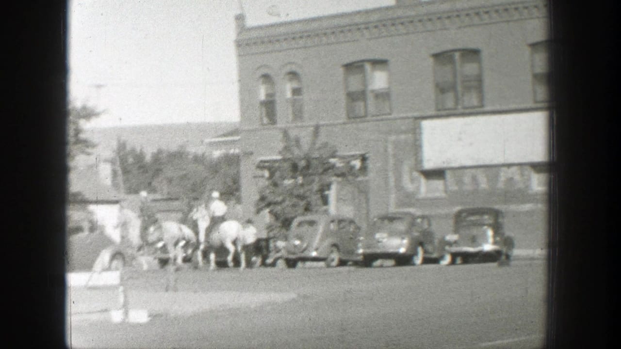 Horse riders and pedestrians near parked cars on a town street.