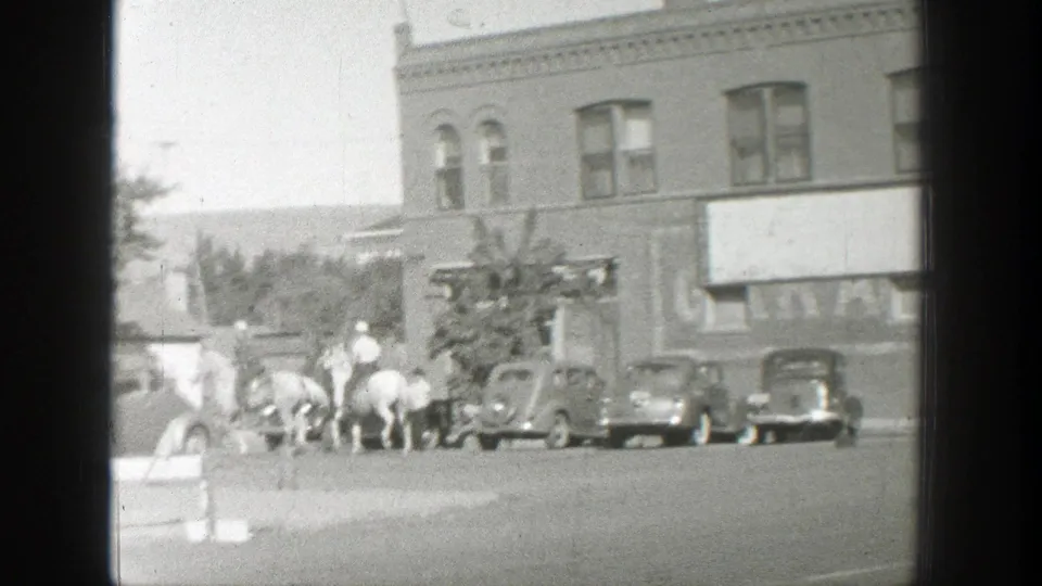 Wyoming street scene with riders, pedestrians, parked late-1930s automobiles, and a brick storefront in soft 8mm focus.