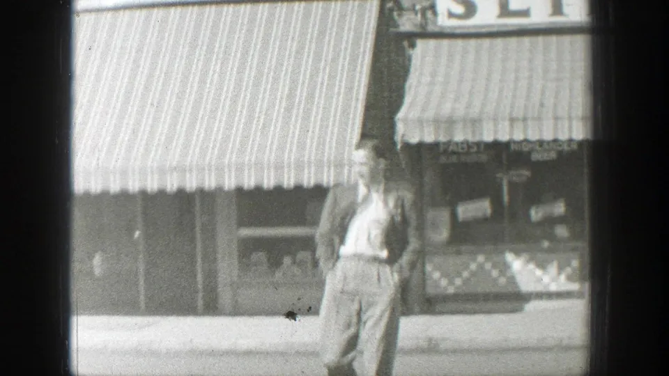 A suited man walks along a Wyoming town sidewalk in front of striped awnings and shop windows in 8mm footage.
