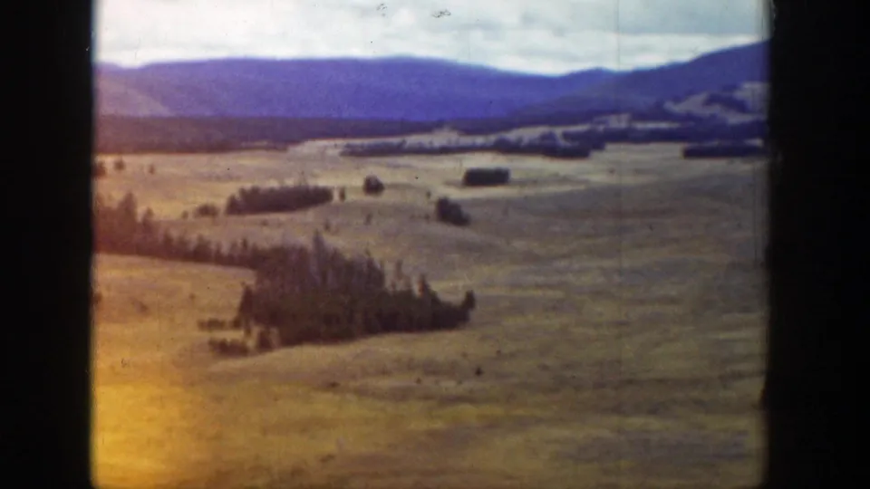 Color-tinted 8mm frame of Wyoming fields, tree lines, and distant mountains under a cloudy sky.