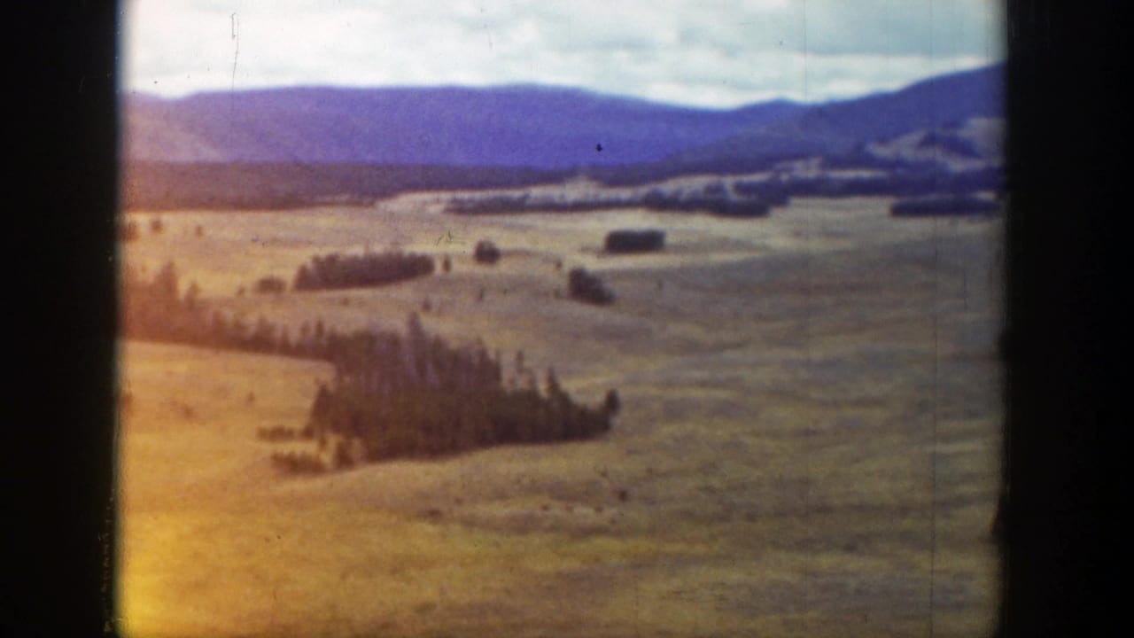 1939 Wyoming 8mm frame showing rolling fields, dark tree clusters, and purple-blue mountains beneath a cloudy sky.