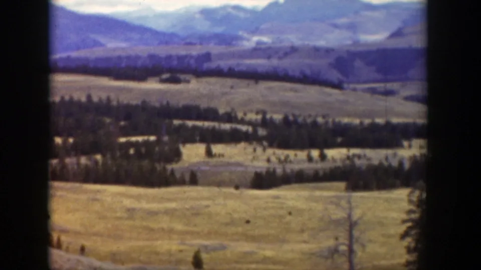 Wyoming high-plains view from 1939 home-movie footage, with yellow hills, evergreen clusters, and distant mountains.