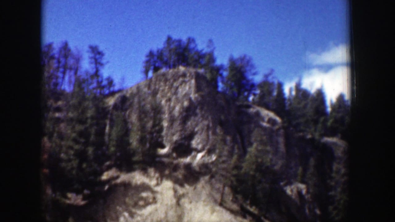 Sunlit craggy cliff with evergreen trees under a blue sky, framed by dark film borders.